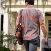 A man in a Tom Beckbe Tidewater Shirt with UPF 30+, carrying a black bag and pulling a suitcase past hanging plants.