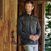 A man in a dark Tom Beckbe Tucker Fleece Quarter Zip stands by a wooden doorway, looking aside with trees behind.