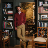Man in a Tom Beckbe Tucker Fleece Quarter Zip and khaki pants leans on a bookshelf in a study with wood chairs.