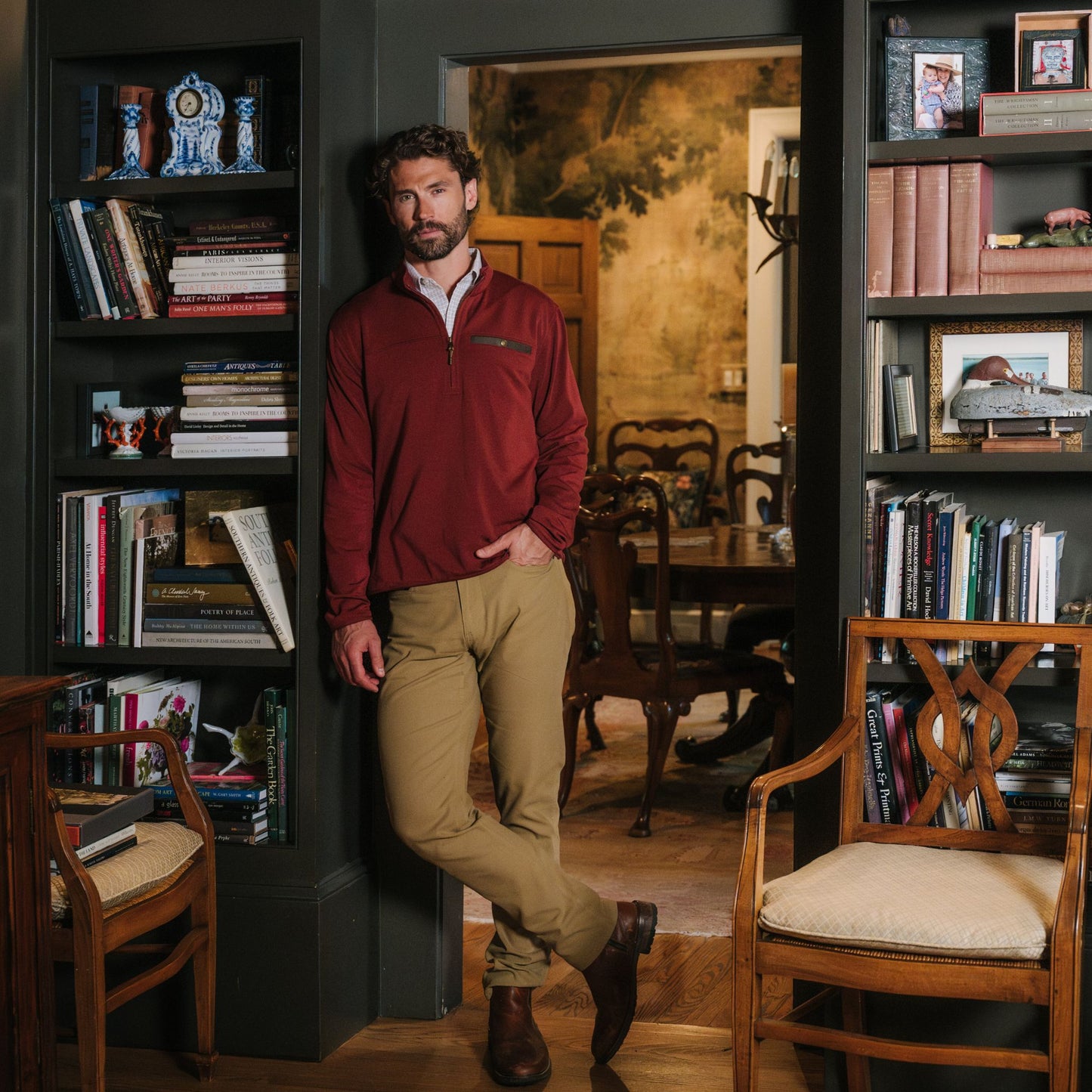 Man in a Tom Beckbe Tucker Fleece Quarter Zip and khaki pants leans on a bookshelf in a study with wood chairs.