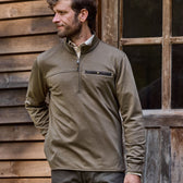 A man stands by a wooden building in a Tom Beckbe Tucker Fleece Quarter Zip, glancing aside with zip and buttoned chest pocket.