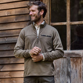 Man in an olive green Tom Beckbe Tucker Fleece Quarter Zip smiles warmly by a wooden wall and window.
