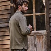 Man in an olive jacket faces a wooden building wearing a Tom Beckbe Tucker Fleece Quarter Zip.