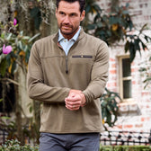 Man in a brown Tom Beckbe Tucker Fleece Quarter Zip stands outside a brick building, blooming plants behind.