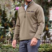 A person in a khaki Tucker Fleece Quarter Zip by Tom Beckbe stands outdoors amid lush greenery.