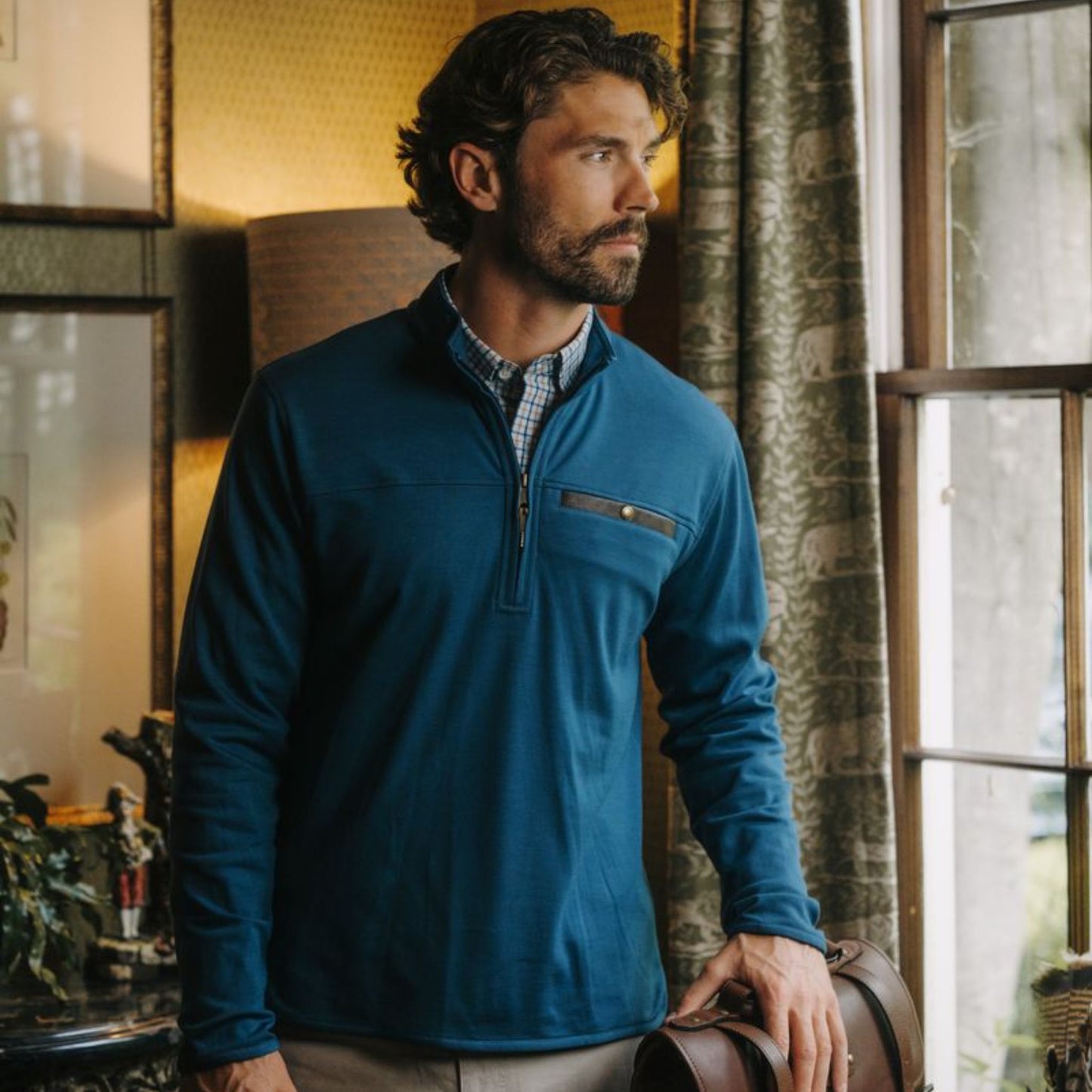 Man with wavy hair in a Tom Beckbe Tucker Fleece Quarter Zip stands by a window, holding a leather bag.