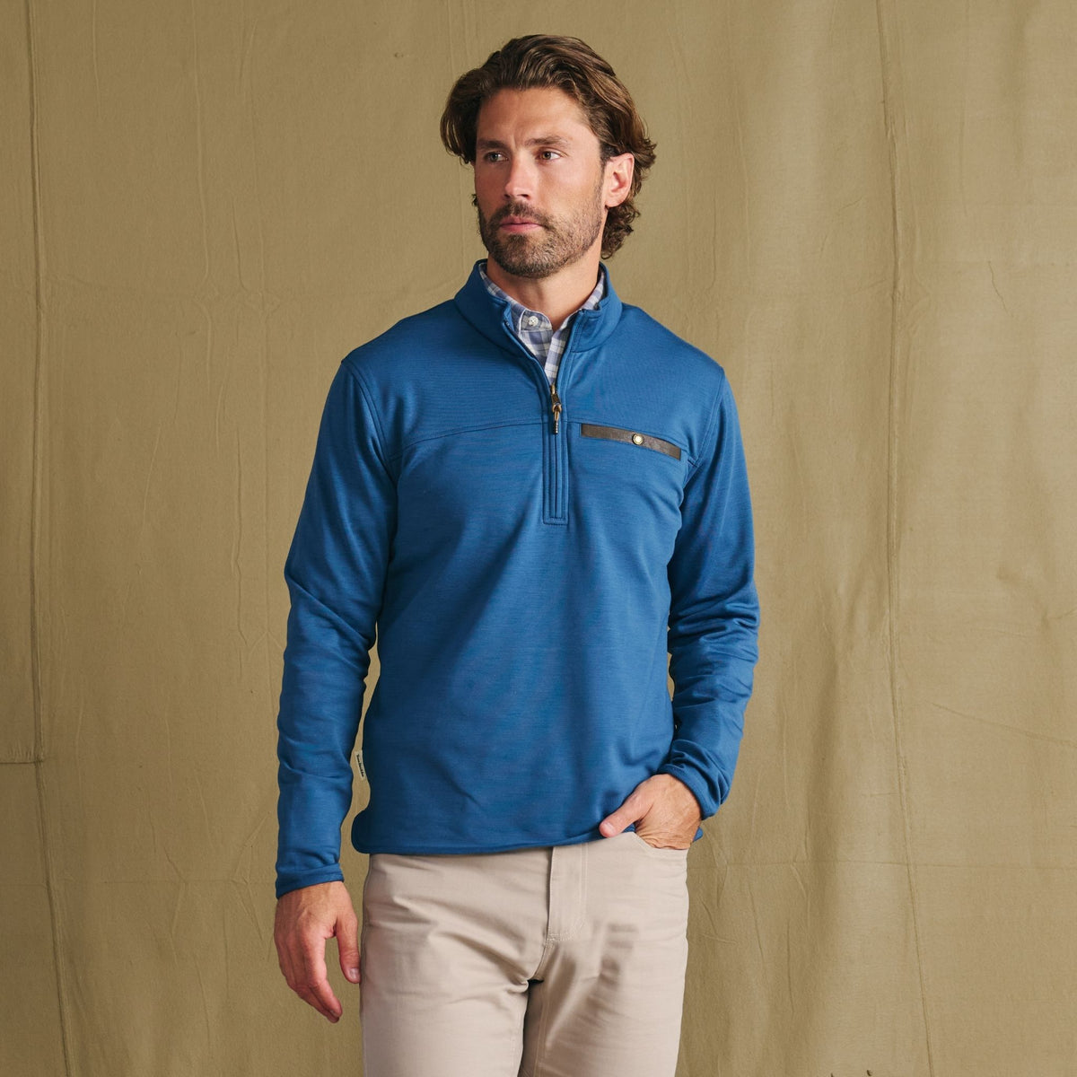 Man in the Tom Beckbe Tucker Fleece Quarter Zip and beige pants stands before a tan backdrop, gazing to the side.