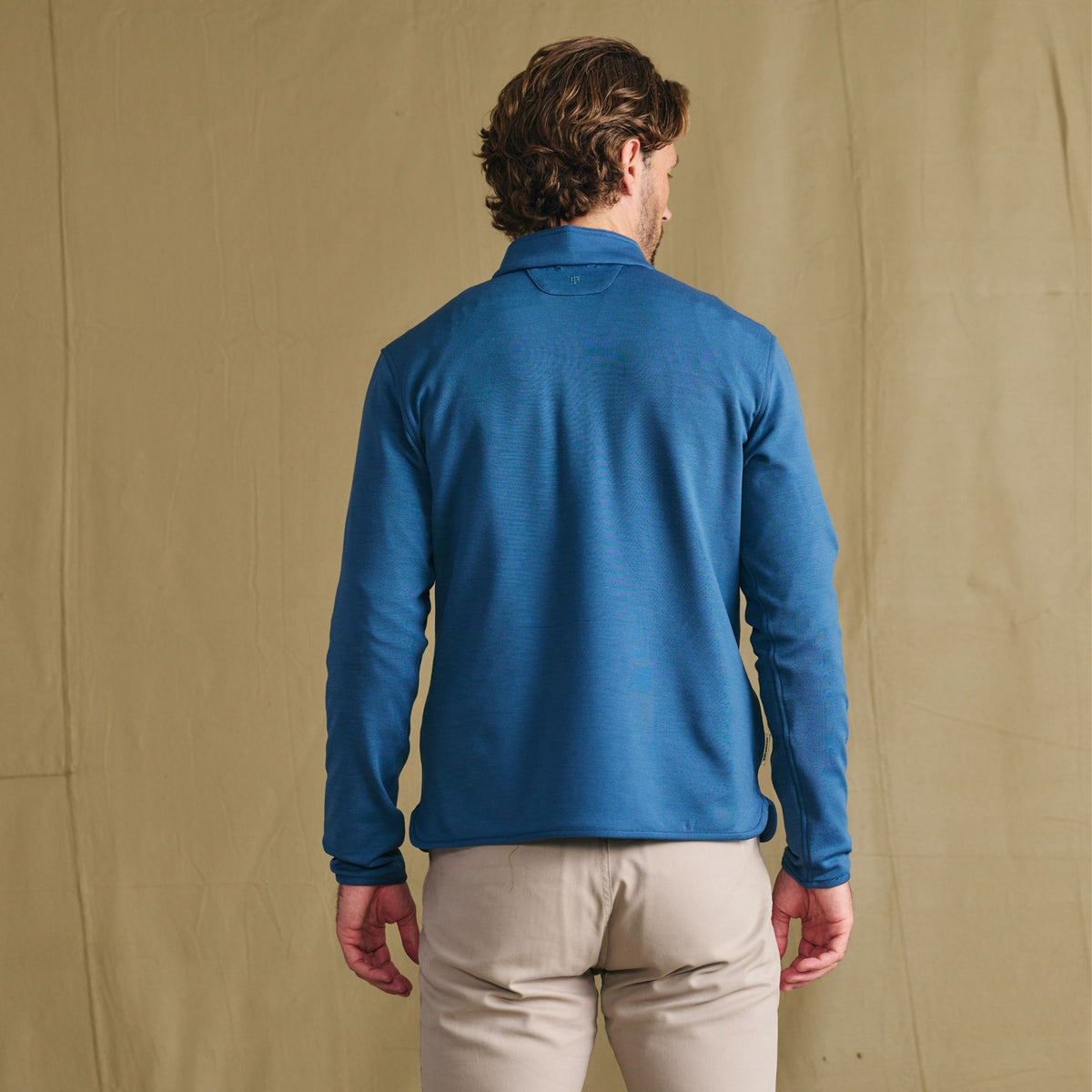 Man facing away, wearing the Tom Beckbe Tucker Fleece Quarter Zip in blue with beige pants against a tan background.