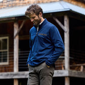 Man in a blue Tom Beckbe Tucker Fleece Quarter Zip, hands in pockets, stands by a rustic wooden building outdoors.