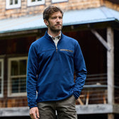 A man in a blue Tom Beckbe Tucker Fleece Quarter Zip stands outdoors by a wooden building, gazing into the distance.