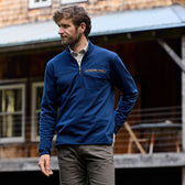 Man in a blue quarter zip and khaki pants stands by a rustic building, showcasing the Tom Beckbe Tucker Fleece.