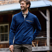 Man in a Tom Beckbe Tucker Fleece Quarter Zip smiles, hands in pockets, by a rustic building with a metal roof.
