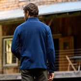 A man wearing a Tom Beckbes Tucker Fleece Quarter Zip gazes at a rustic wooden building with a metal roof outdoors.