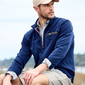 A man in a Tom Beckbe Tucker Fleece Quarter Zip, checkered shirt, khaki shorts, and beige cap sits outdoors, gazing sideways.