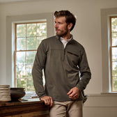 A man in a green Tom Beckbe Tucker Fleece Quarter Zip stands indoors by a table, glancing sideways as sunlight pours in.