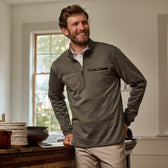 A man in a Tom Beckbe Tucker Fleece Quarter Zip stands by a table with dishes, smiling indoors near a window.