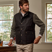 Man in a Tom Beckbe Tucker Fleece Quarter Zip and dark vest leans on a wooden table in a room with large windows.