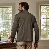 A man in a Tom Beckbe Tucker Fleece Quarter Zip stands by a table, facing windows with green trees outside.