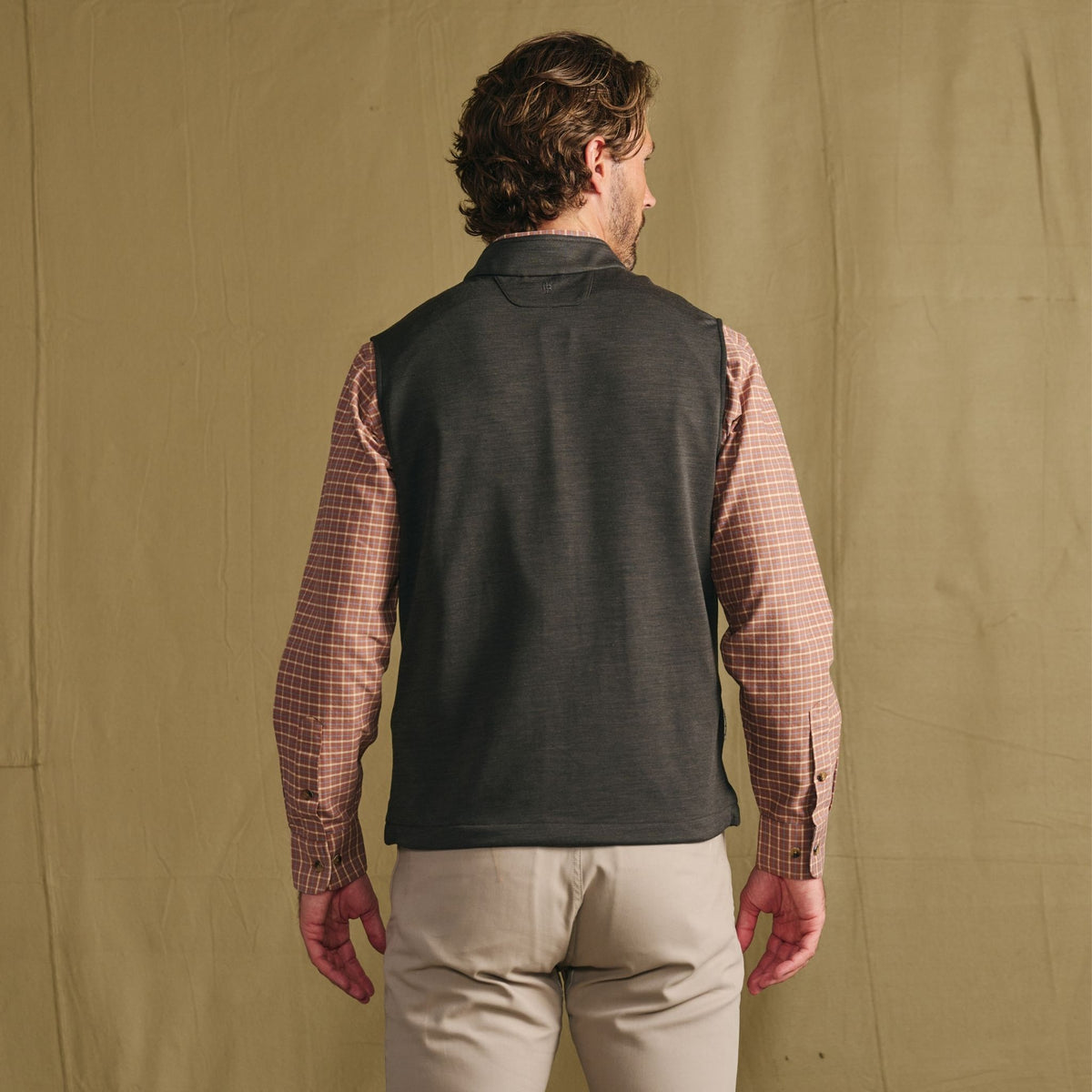 Man facing away in a Tom Beckbe Tucker Fleece Vest over plaid shirt, light pants, against a beige fabric backdrop.