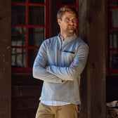 A man in a light gray Tom Beckbe Tucker Fleece Quarter Zip stands arms crossed by a wooden building with red-trimmed windows.