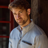 Light brown-haired man with beard in Tom Beckbes Tucker Fleece Quarter Zip, standing before a dark wooden wall.
