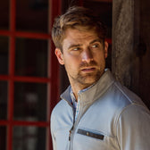 Man with light brown hair and beard in a Tom Beckbe Tucker Fleece Quarter Zip stands by a red-windowed building, looking aside.