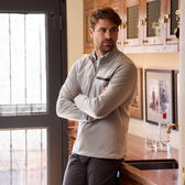 Man in a light gray Tom Beckbe Tucker Fleece Quarter Zip stands with arms crossed by a counter, pictures behind him.