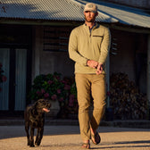 A man in a beige outfit and flip-flops, with a black dog, wears a Tom Beckbe Tucker Fleece Quarter Zip near a rustic building.
