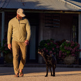 A man in casual attire and a Tom Beckbe Tucker Fleece Quarter Zip walks with a black dog by a building with plants.