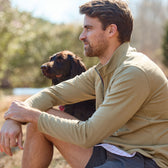 A man in a Tom Beckbe Tucker Fleece Quarter Zip sits outside with his brown dog, both gazing into the distance.