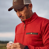 Man in a red jacket, baseball cap looks down, holding an object. Blurred background. Tucker Fleece Quarter Zip by Tom Beckbe.
