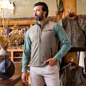 A man in a beige vest and green plaid shirt stands in a rustic clothing store with jackets and hats displayed around him.