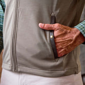 A person’s hand rests in the pocket of a light brown zip-up vest, worn over a green plaid shirt.