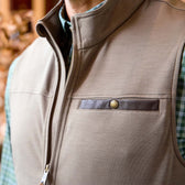 Close-up of a person wearing a tan zip-up vest with a leather-trimmed chest pocket over a green plaid shirt.