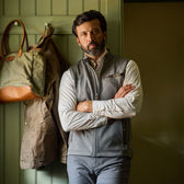 Man with beard wearing a gray vest and striped shirt stands with arms crossed in front of a coat rack with bags and a jacket.