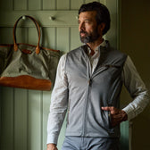 Man wearing a gray zip-up vest and plaid shirt stands indoors next to a wall with a hanging green and brown bag.