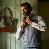 Man zipping up a gray vest over a checked shirt, standing indoors near a green wall with a tote bag hanging on a hook.