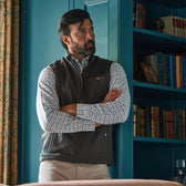 Man with a beard stands with arms crossed, wearing a vest and checked shirt, leaning against a blue bookcase.