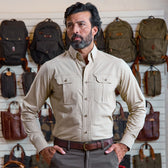 A man wears the Tom Beckbe Uplander Shirt, standing by a wall filled with backpacks and bags.