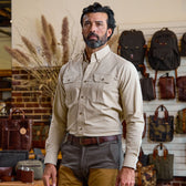A man indoors wears the Tom Beckbe Uplander Shirt in tan cotton oxford and brown pants; leather bags are behind him.