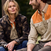 Two people sit outdoors—one in a camouflage jacket, the other in a Tom Beckbe Uplander Shirt with an orange yoke.