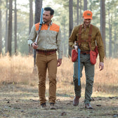 Two men in outdoor attire, wearing Uplander Shirts by Tom Beckbe and safety orange caps, walk through the woods with shotguns.