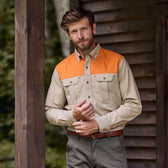 A man in a Tom Beckbe Uplander Shirt stands by a rustic wooden post with greenery in the background.