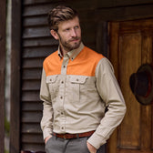 Bearded man in a Tom Beckbe Uplander Shirt, orange and beige, hands in pockets by wooden building exterior.