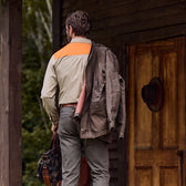 A man in a tan Tom Beckbe Uplander Shirt with an orange panel holds a jacket and bag near a cabin outdoors, facing a wooden door.