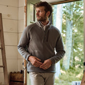 A man in a gray Tom Beckbe Warren Fleece Quarter Zip stands in a doorway, gazing at trees in the soft natural light.