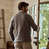 A man in a Tom Beckbe Warren Fleece Quarter Zip opens a wooden door to a bright outdoor scene; the rustic interior has wood furniture.