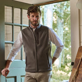 A man in a Tom Beckbe Warren Fleece Vest over a plaid shirt stands at an open door, hand on hip, with a forest backdrop.
