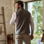 A man in a gray Tom Beckbe Warren Fleece Vest and plaid shirt gazes at the sunny woods through an open door.
