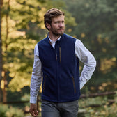A man wearing a Tom Beckbe Warren Fleece Vest and a checked shirt walks outdoors with trees in the background.